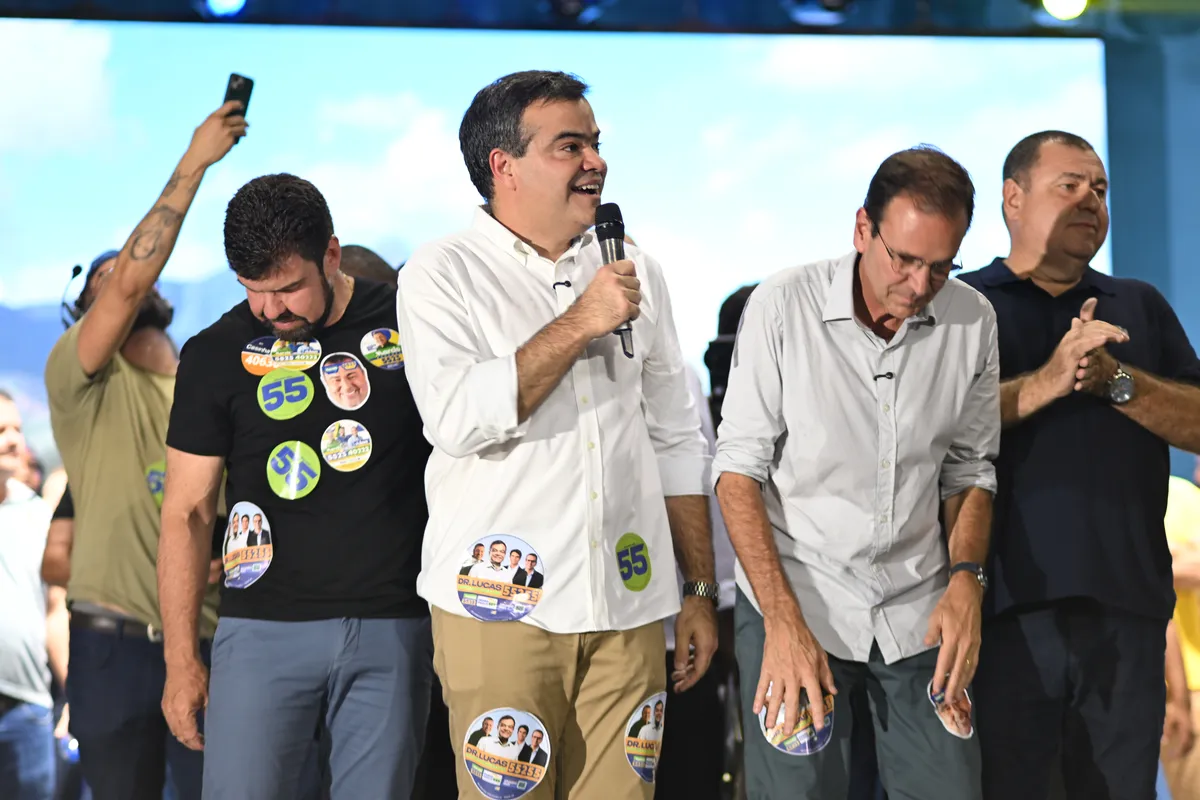 Dr. Lucas 55255 no palco, de microfone na mão, sorrindo ao lado de outros candidatos da coligação