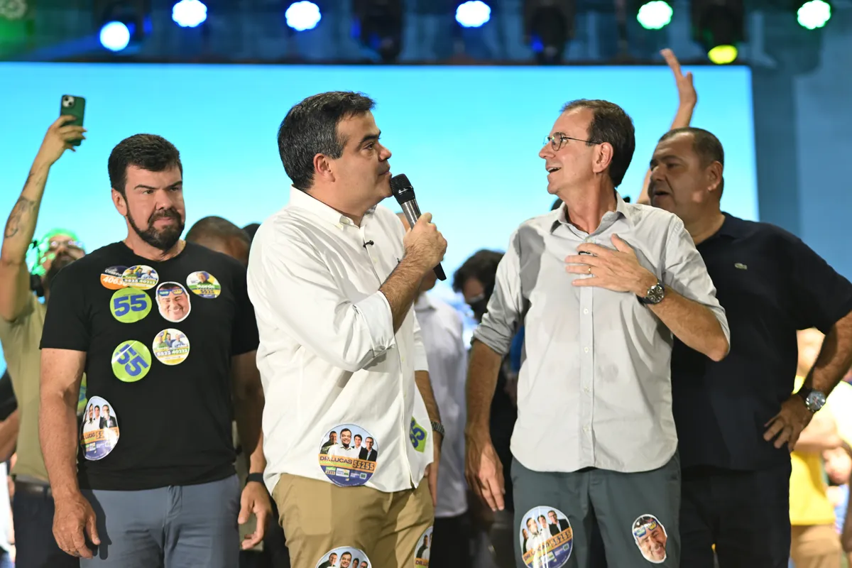 Dr. Lucas 55255 fala ao microfone no palco enquanto conversa com um candidato ao lado