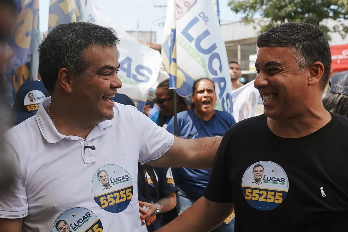 Dr. Lucas 55255 cumprimenta um apoiador com adesivo 55255, cercado de bandeiras, em Rio das Ostras