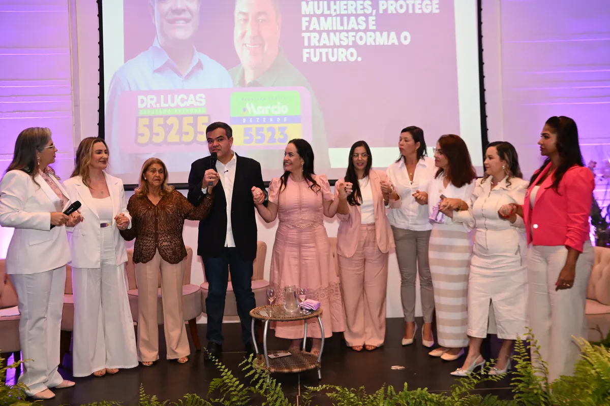 Dr. Lucas 55255 de braços dados com as mulheres no palco, com o telão da campanha ao fundo