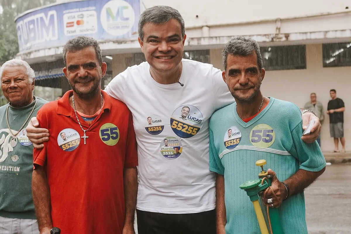 Dr. Lucas 55255 abraçado a três apoiadores na rua, sob chuva fina, em Conceição de Macabu