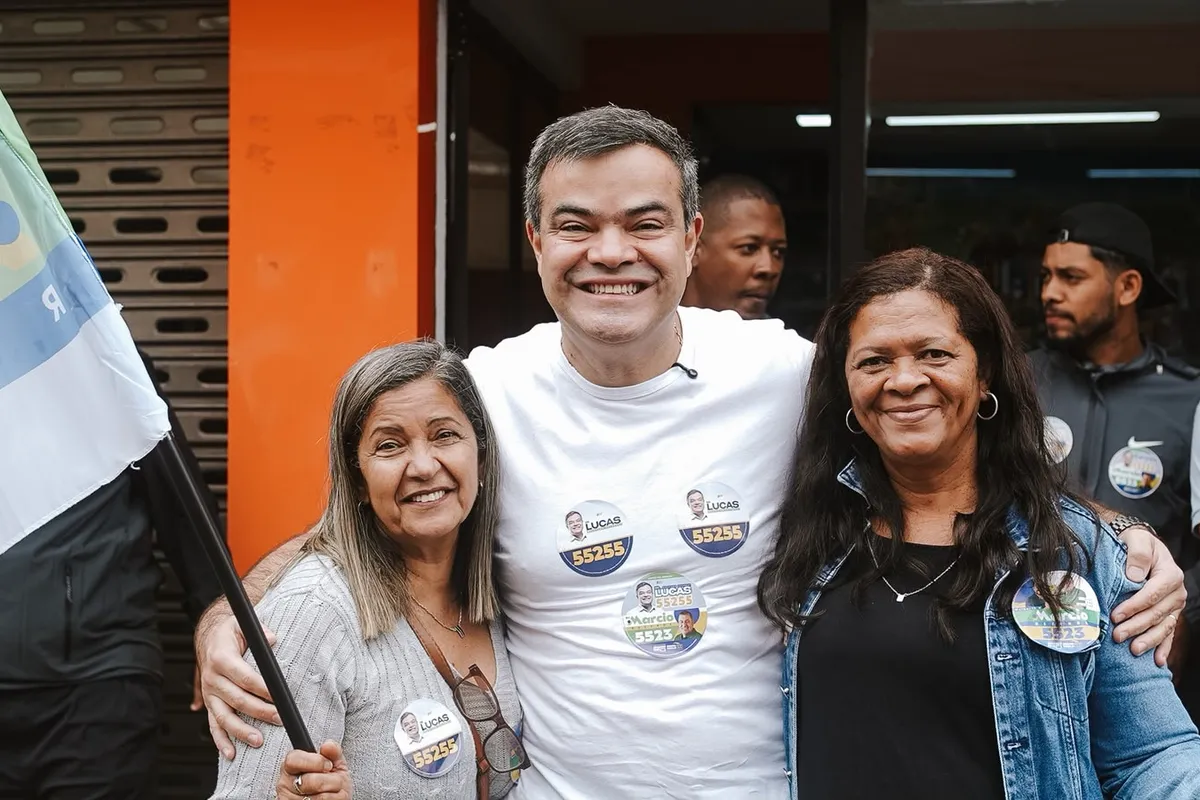 Dr. Lucas 55255 entre duas apoiadoras, com bandeira da campanha ao fundo, em Conceição de Macabu